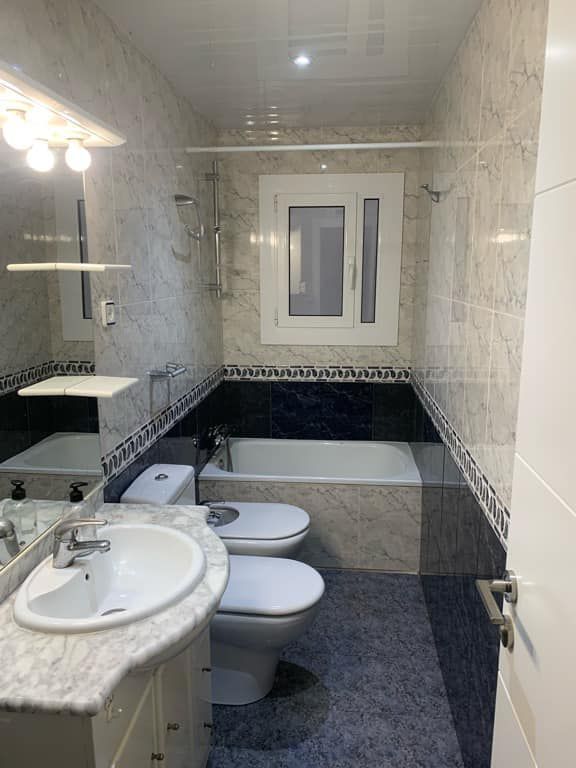 Baño con lavabo, inodoro, bidé y bañera blancos. Paredes y suelo de azulejos azules y blancos.