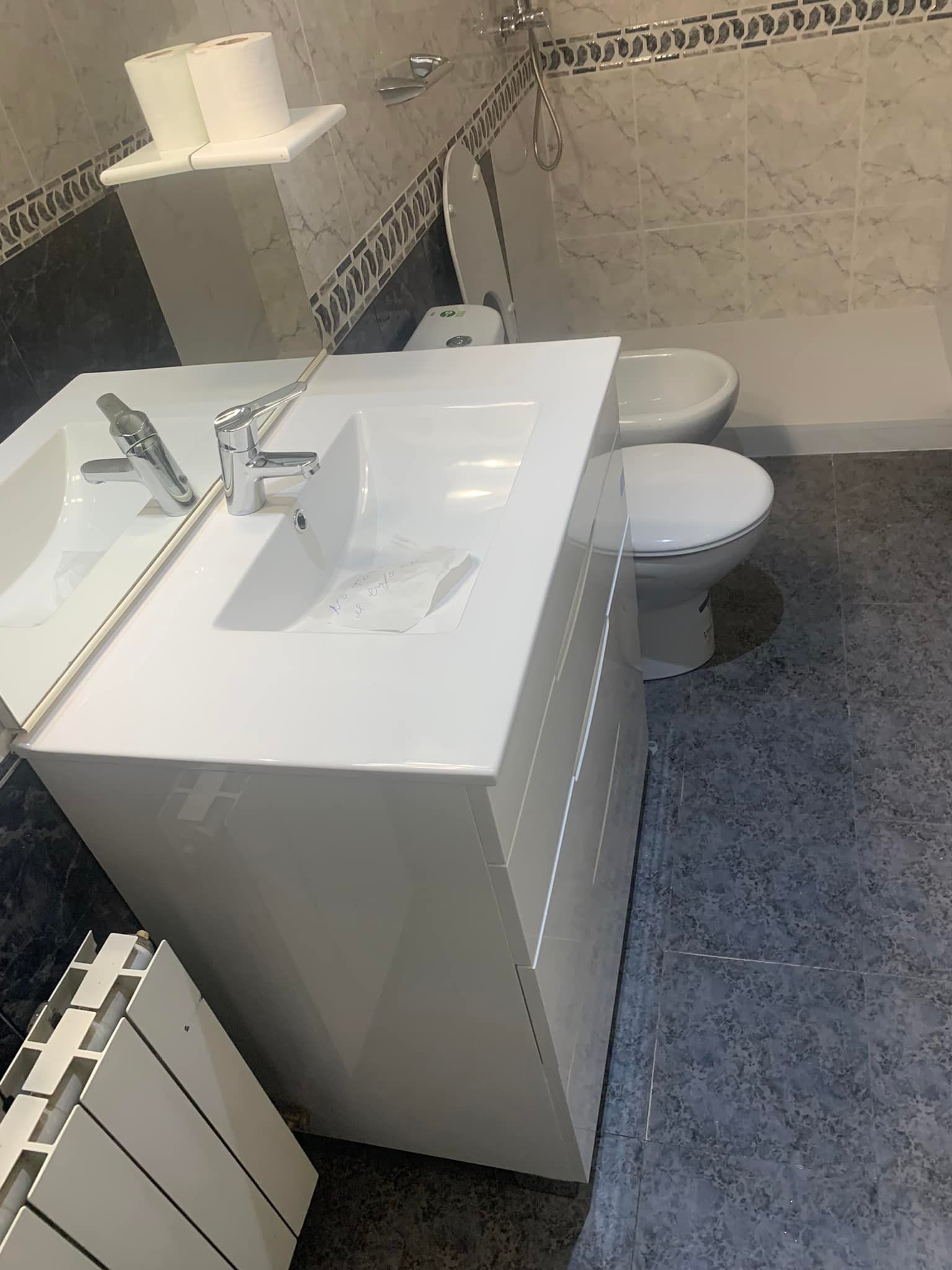 Baño con lavabo y tocador blancos, inodoro y bidé contra paredes de azulejos y suelo de color gris azulado.