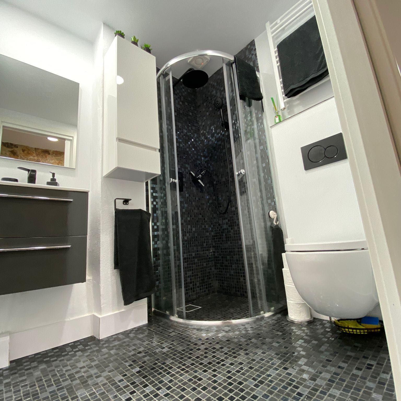 Baño con azulejos negros y grises, ducha, inodoro y tocador.