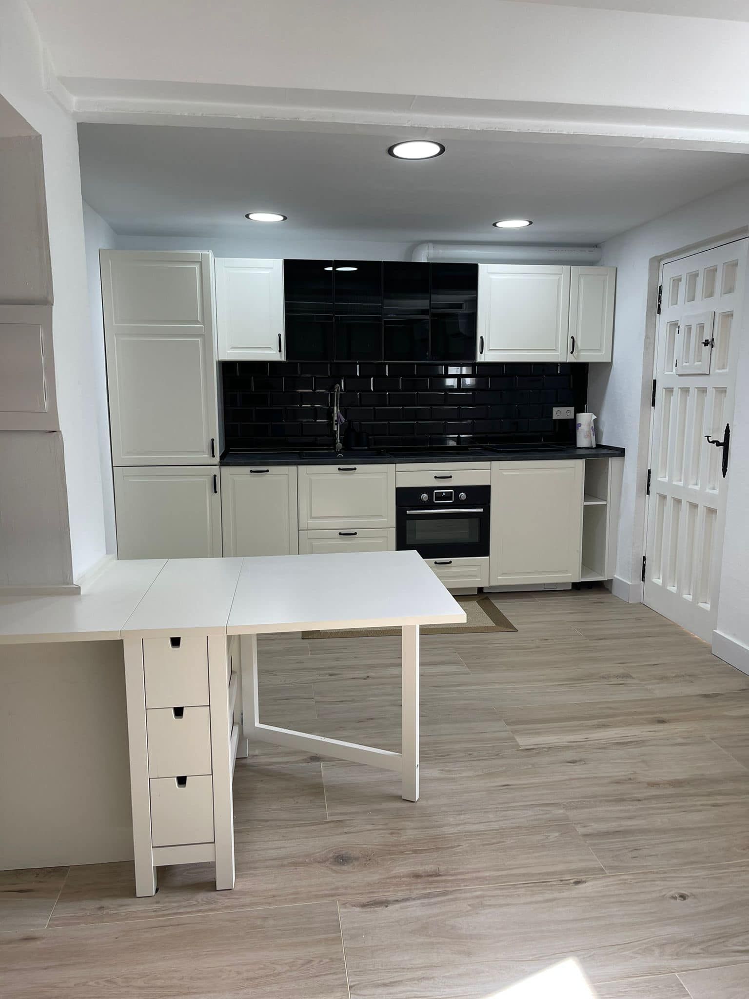 Cocina blanca con salpicadero negro y mesa plegable. Suelos y armarios de color claro.