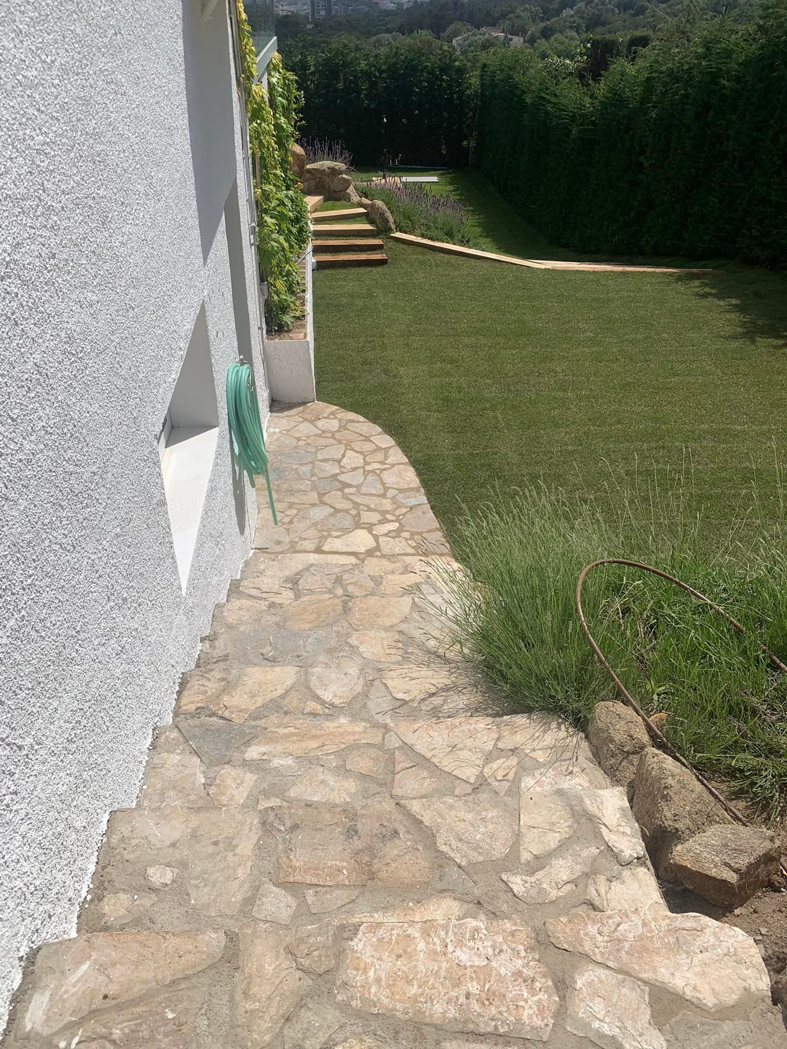 Unos escalones de piedra conducen a un patio cubierto de hierba, delimitado por un seto, junto a un edificio blanco texturizado.