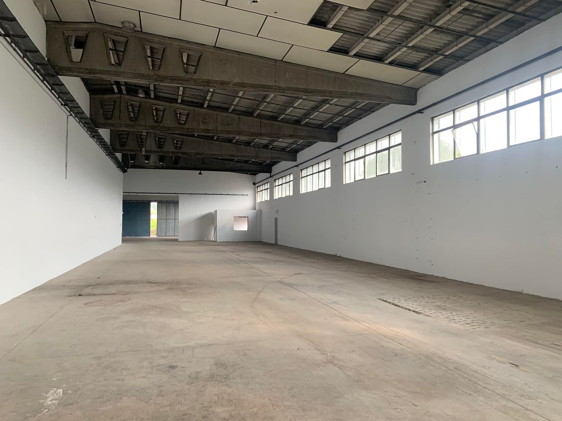 Nave industrial vacía con suelo de hormigón, paredes blancas y ventanas.