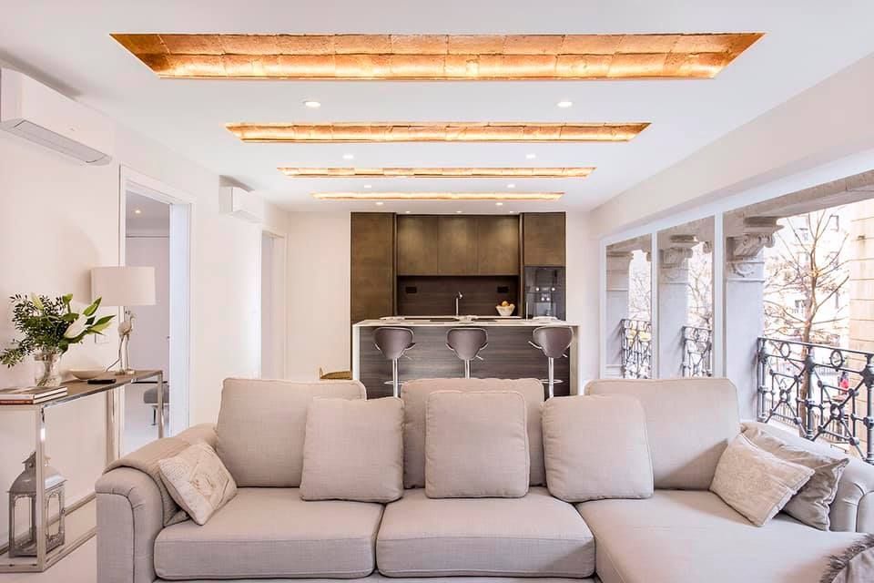Sala de estar con sofá de color claro, cocina abierta al fondo y paneles decorativos en el techo.