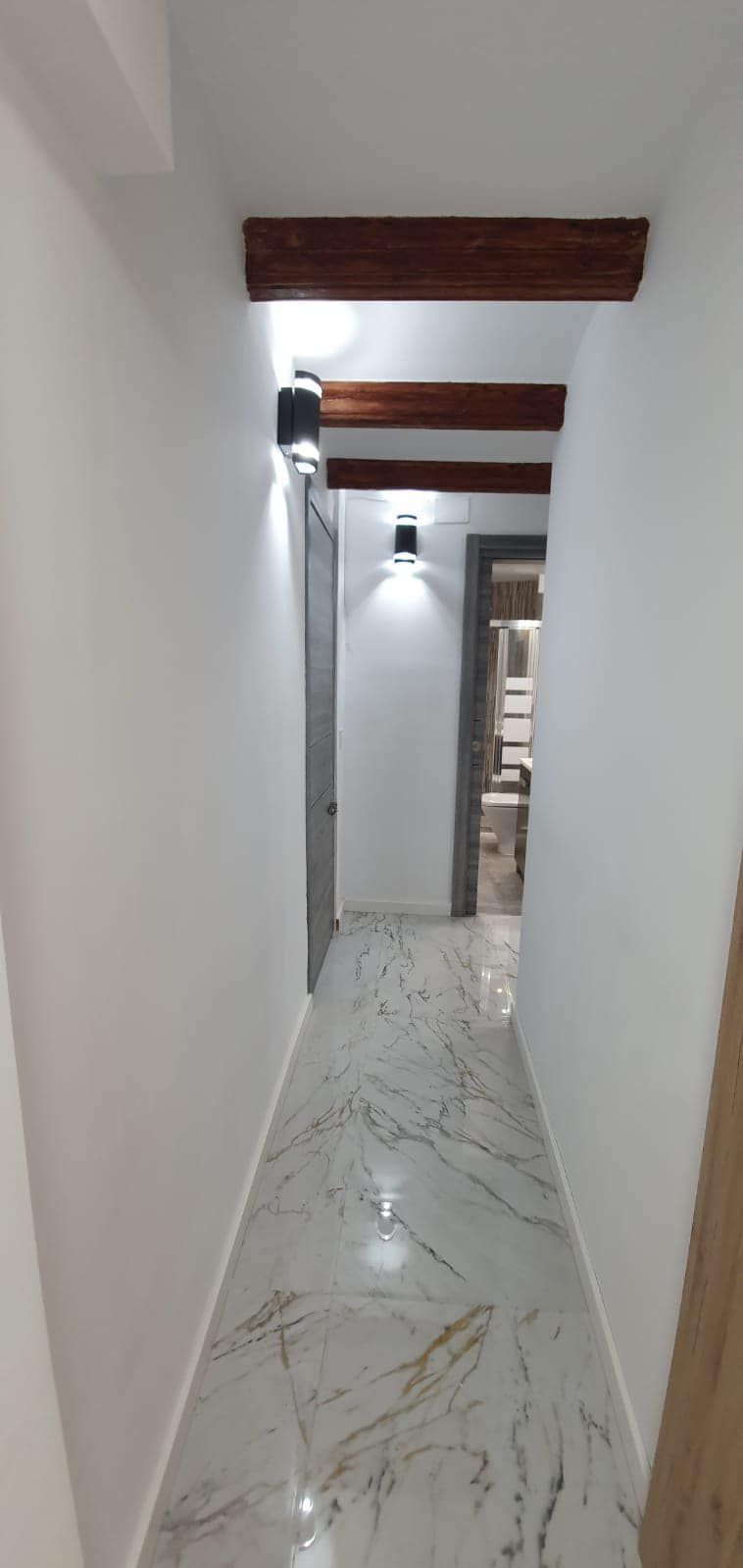 Un pasillo con suelos de mármol, paredes blancas, vigas de madera e iluminación empotrada.