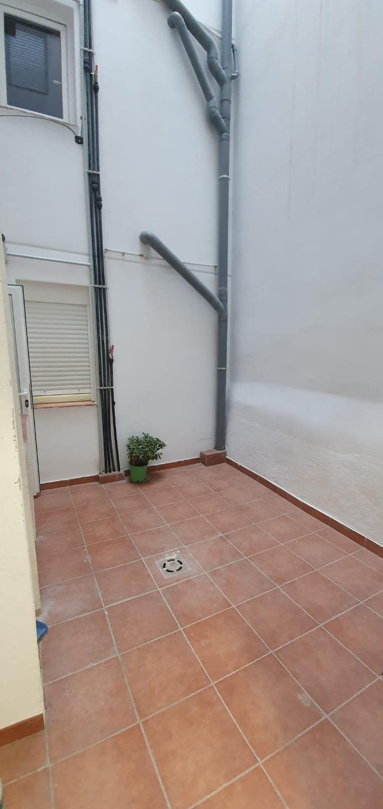 Pequeño patio exterior cerrado con baldosas de terracota y una pequeña maceta. Tuberías grises recorren las paredes blancas.