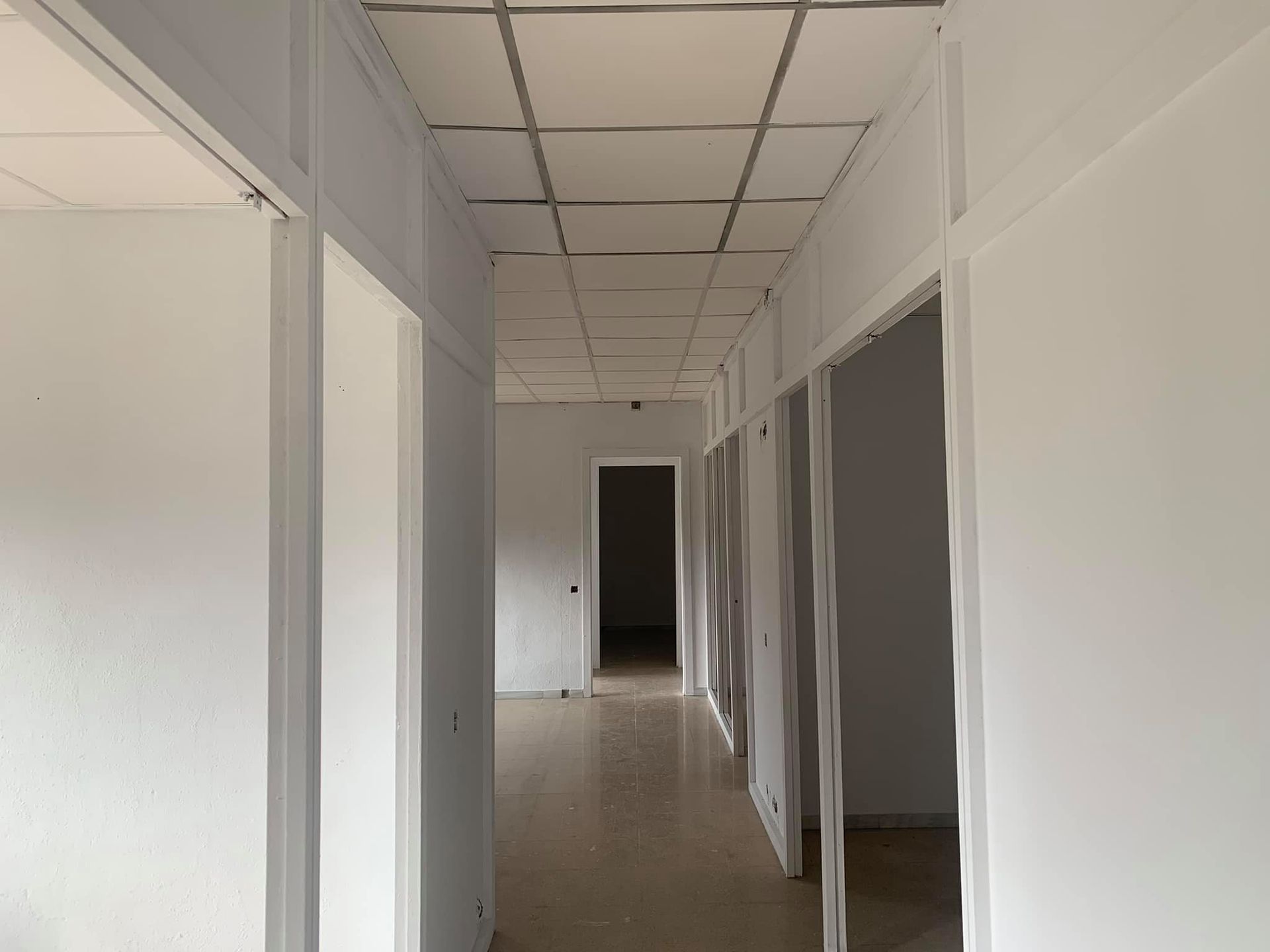 Pasillo blanco con múltiples puertas abiertas y techo suspendido.