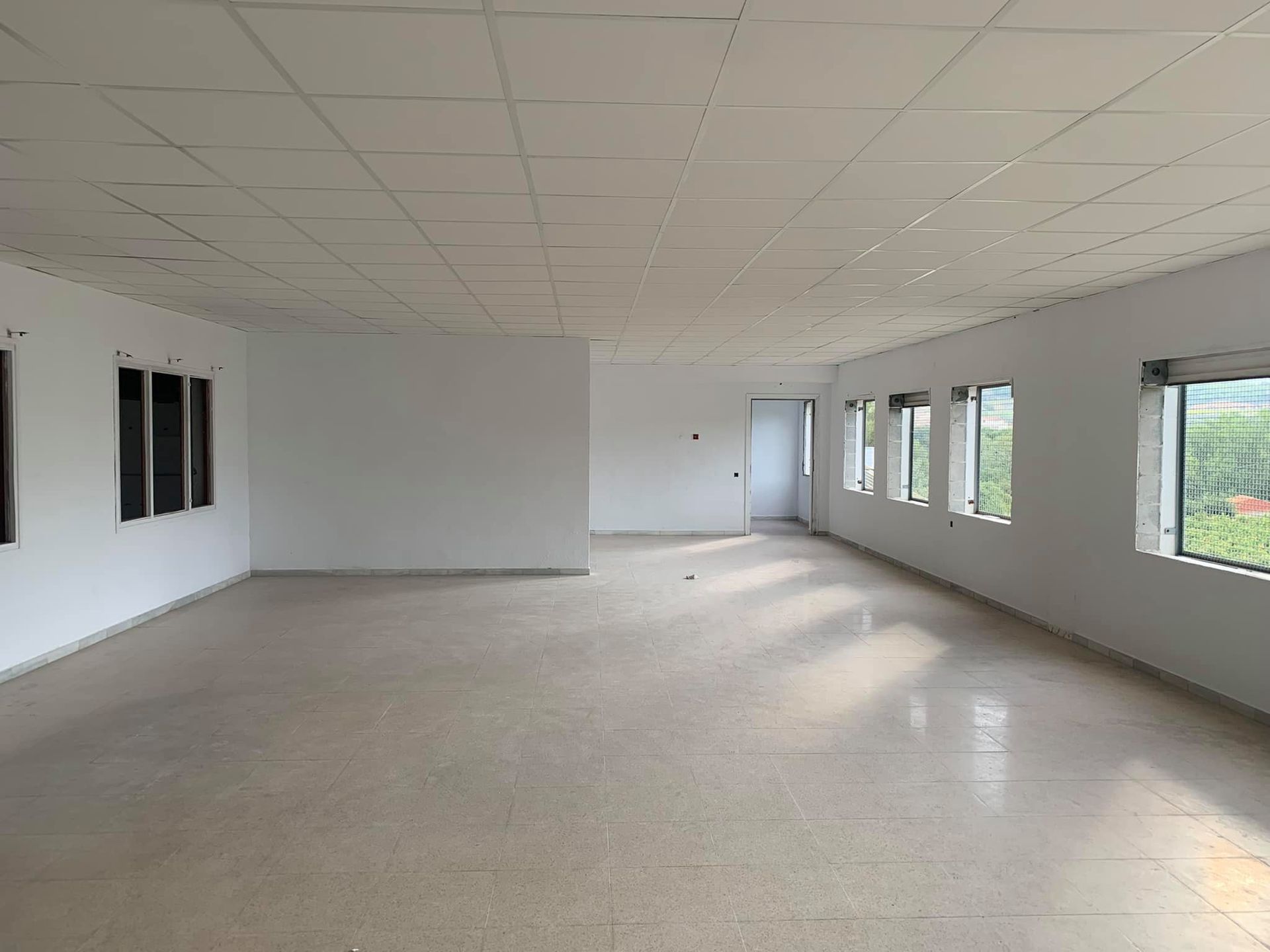 Espacio de oficina vacío y abierto con paredes blancas, ventanas y piso de baldosas.