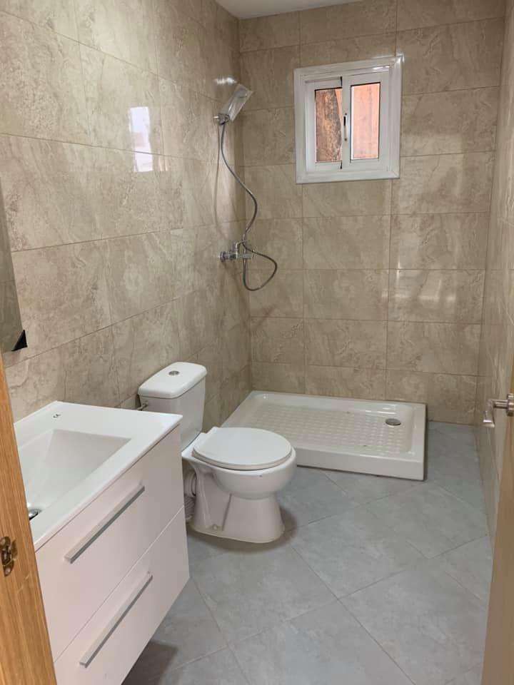 Baño con lavabo blanco, inodoro, ducha y una pequeña ventana. Paredes de azulejos beige y suelo gris.