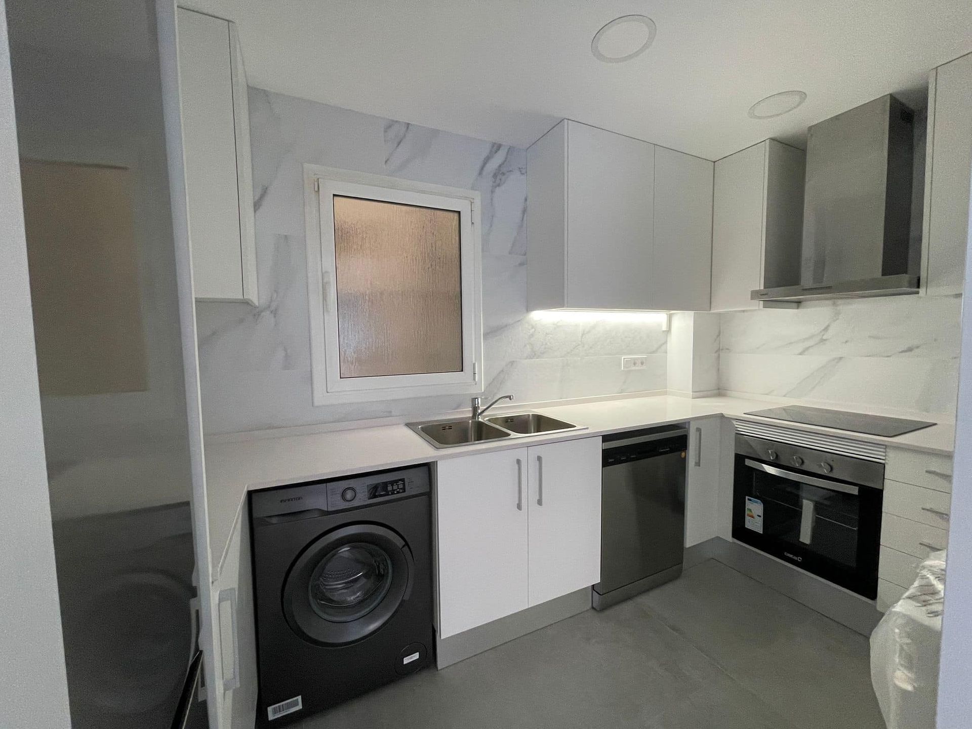 Cocina blanca moderna con electrodomésticos de acero inoxidable, lavadora negra y salpicadero con apariencia de mármol.