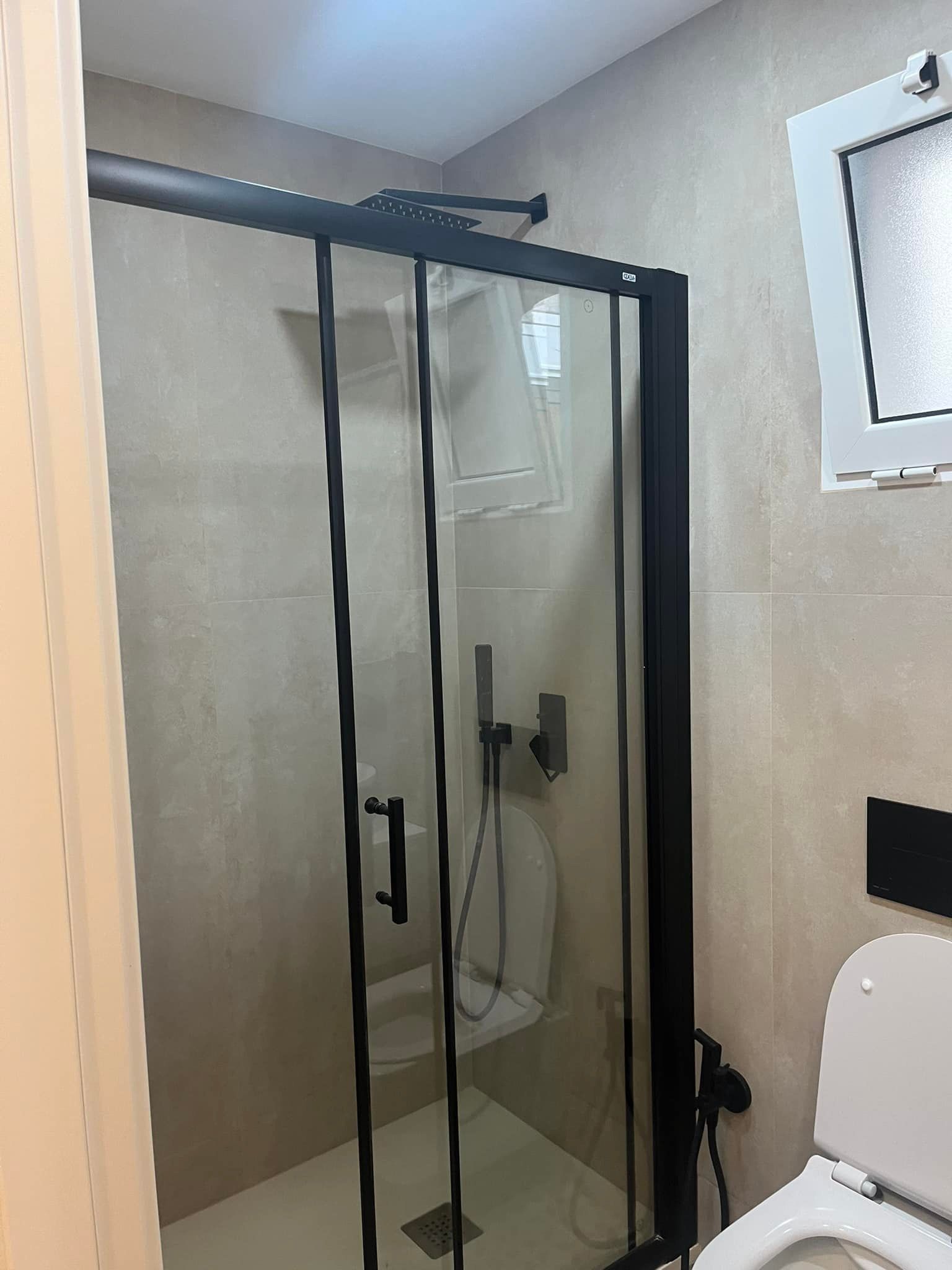 Mampara de ducha con marco negro en un baño con azulejos beige y un inodoro blanco.