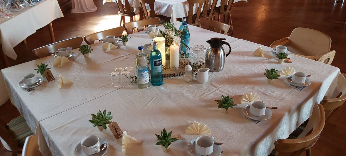 Ein für eine Zusammenkunft gedeckter Tisch mit Kaffeeservice, Kerzen und dekorativen Pflanzen auf einer weißen Tischdecke.