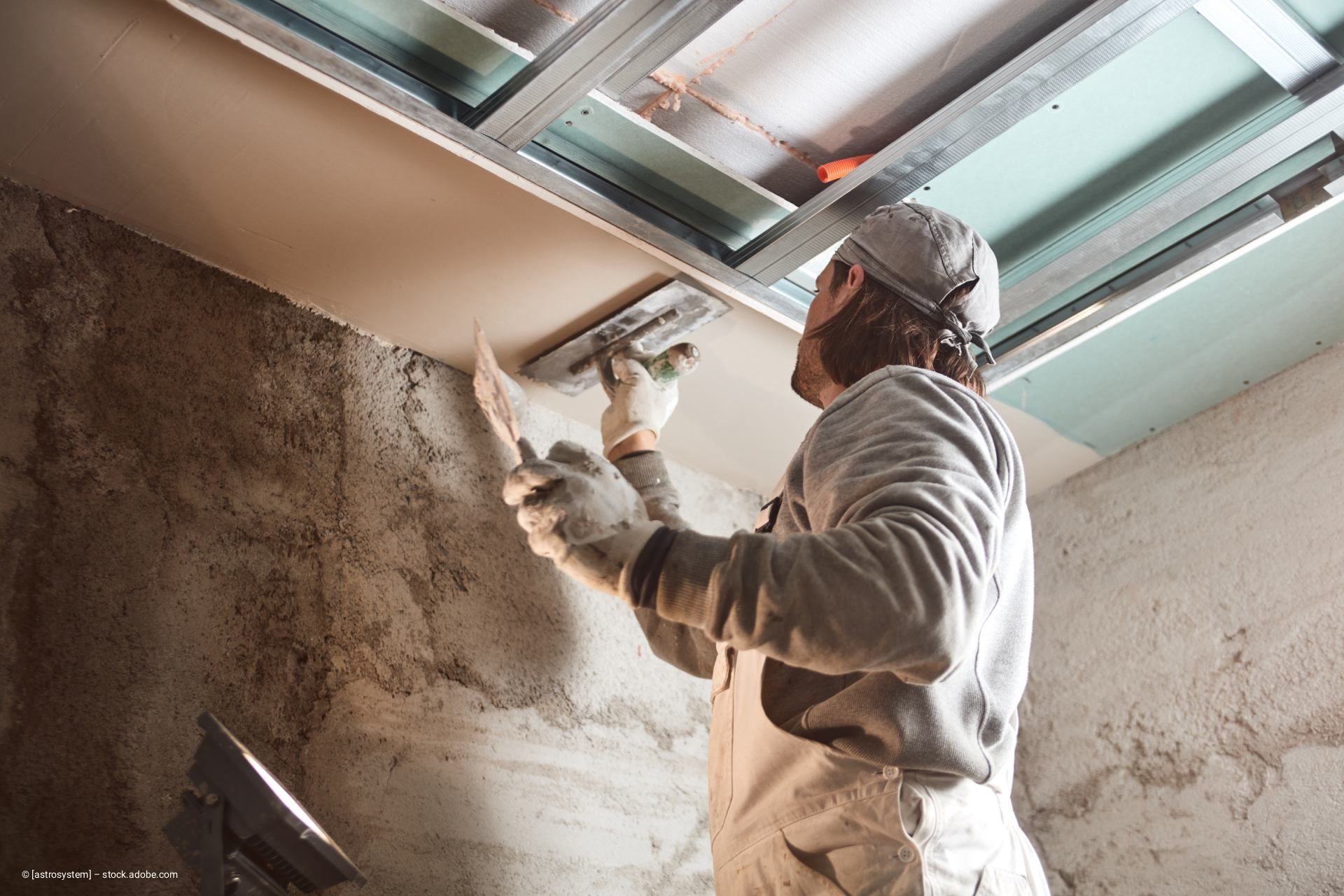 Ein Bauarbeiter verputzt eine Decke; er trägt Handschuhe und benutzt eine Kelle und Glättungswerkzeuge auf einer Baustelle.
