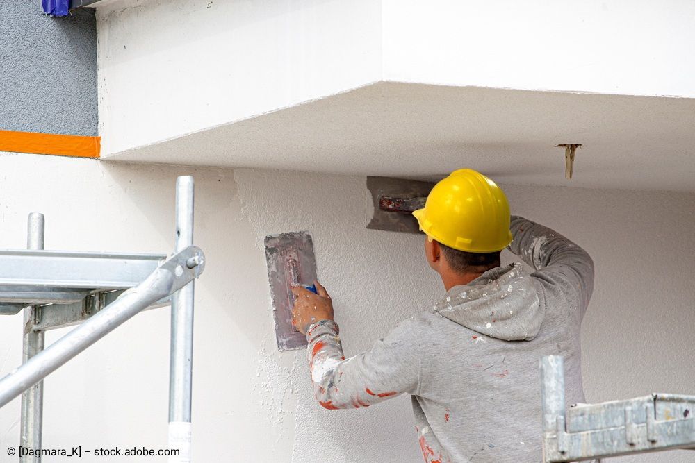 Ein Bauarbeiter mit gelbem Schutzhelm verputzt mit einer Kelle eine weiße Wand.