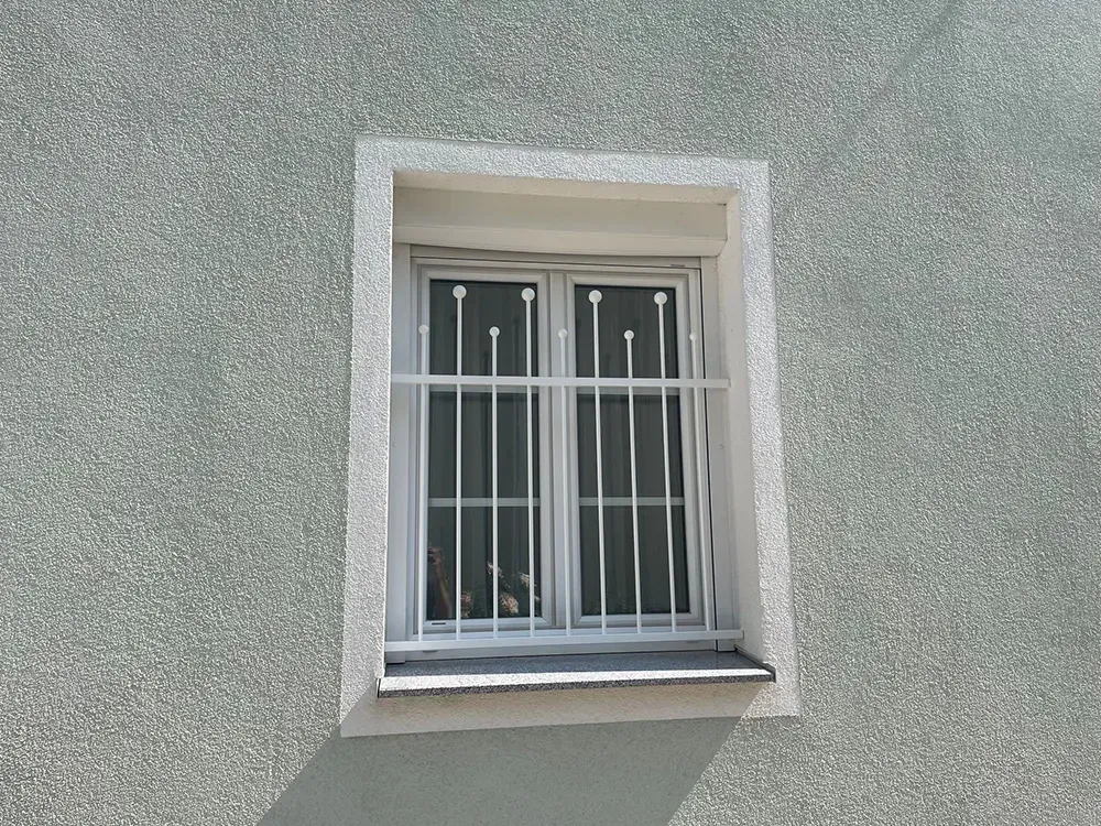 An einer grauen Wand befindet sich ein weißes Fenster mit Gittern.