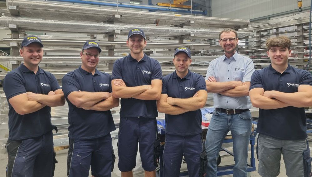 Sechs Personen in Marineuniformen und Freizeitkleidung stehen mit verschränkten Armen in einer Industriehalle.