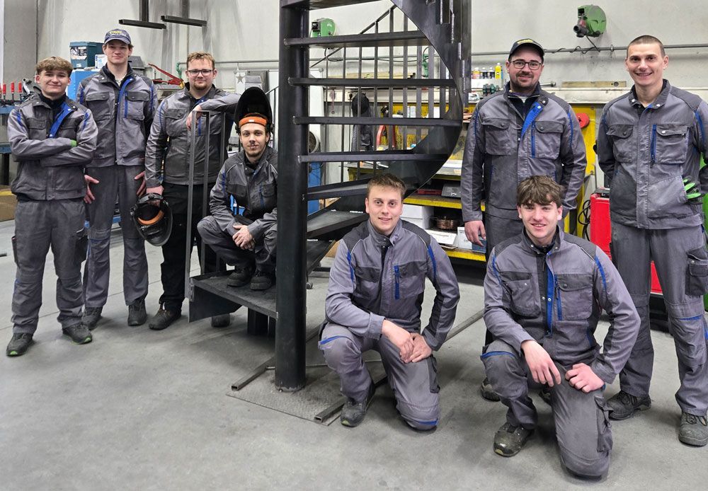 Eine Gruppe von Arbeitern in grauen Uniformen posiert neben einer Metalltreppe in einer Industriewerkstatt.