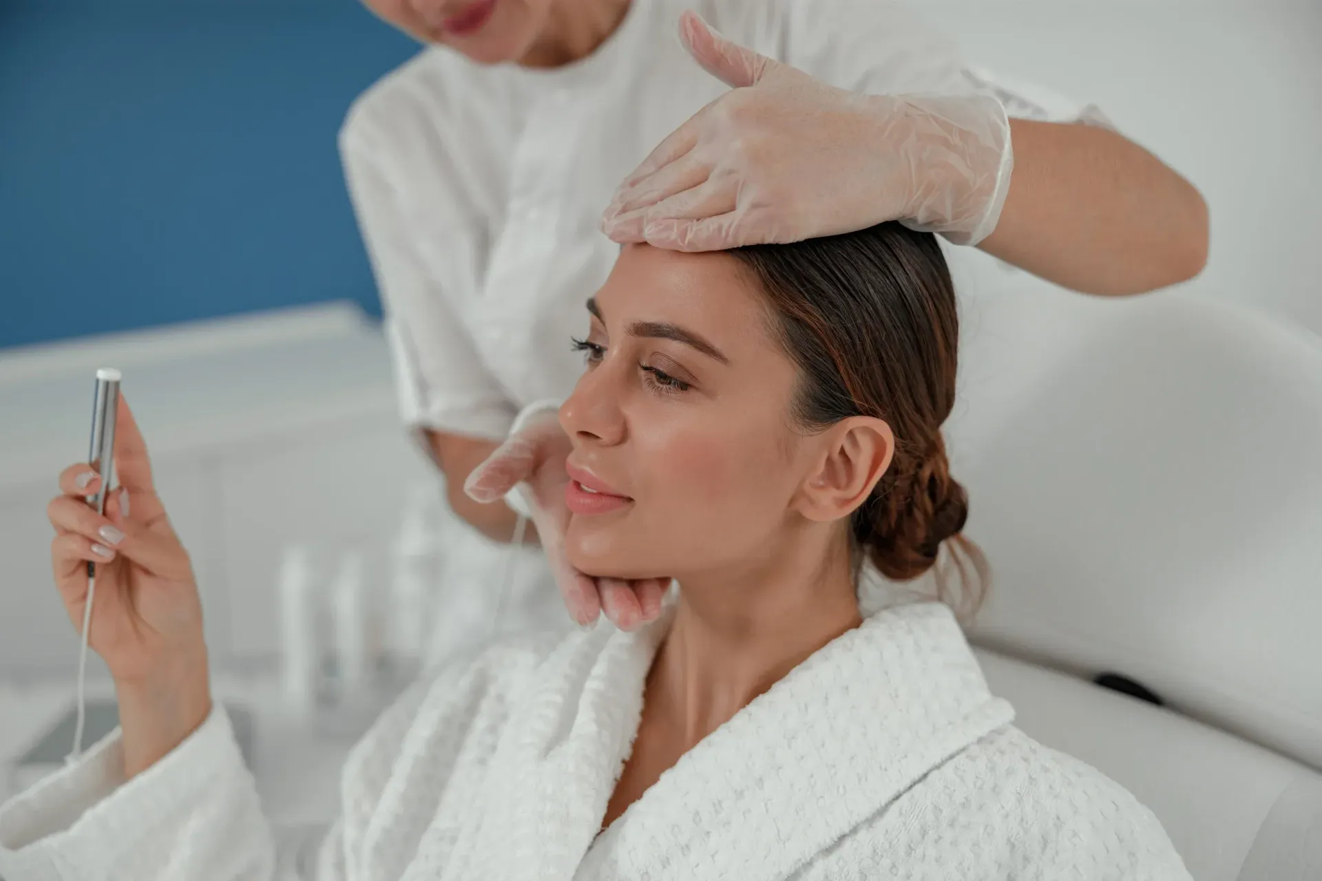 Un profesional del spa, con guantes, aplica un tratamiento facial a un paciente mientras sostiene un dispositivo de mano.