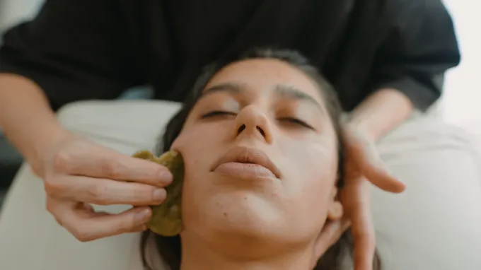 Primer plano de una persona recibiendo un masaje facial con una herramienta gua sha verde sobre una camilla de tratamiento.