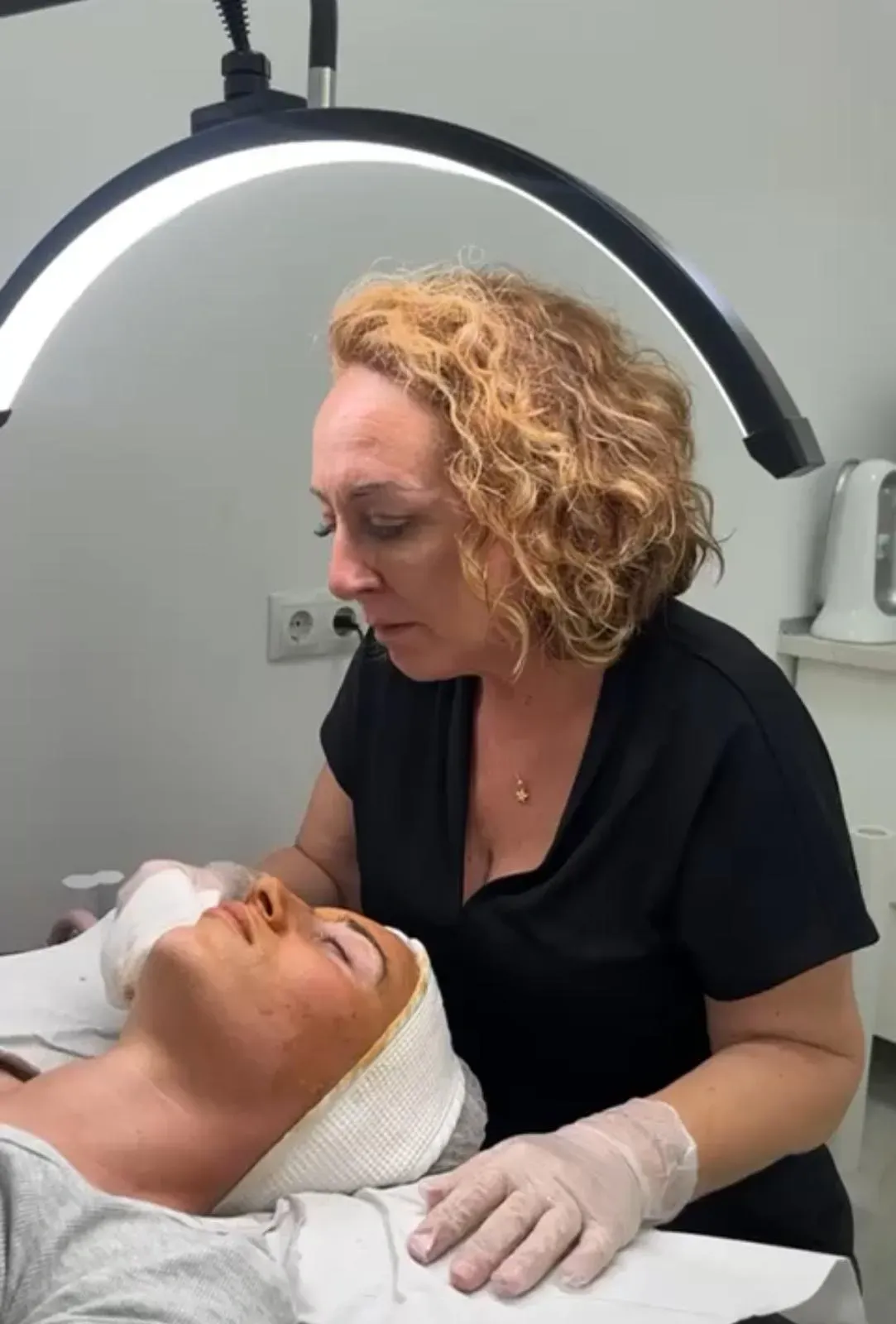 En una clínica, un profesional realiza un tratamiento facial a un cliente que está tumbado bajo una luz anular circular.