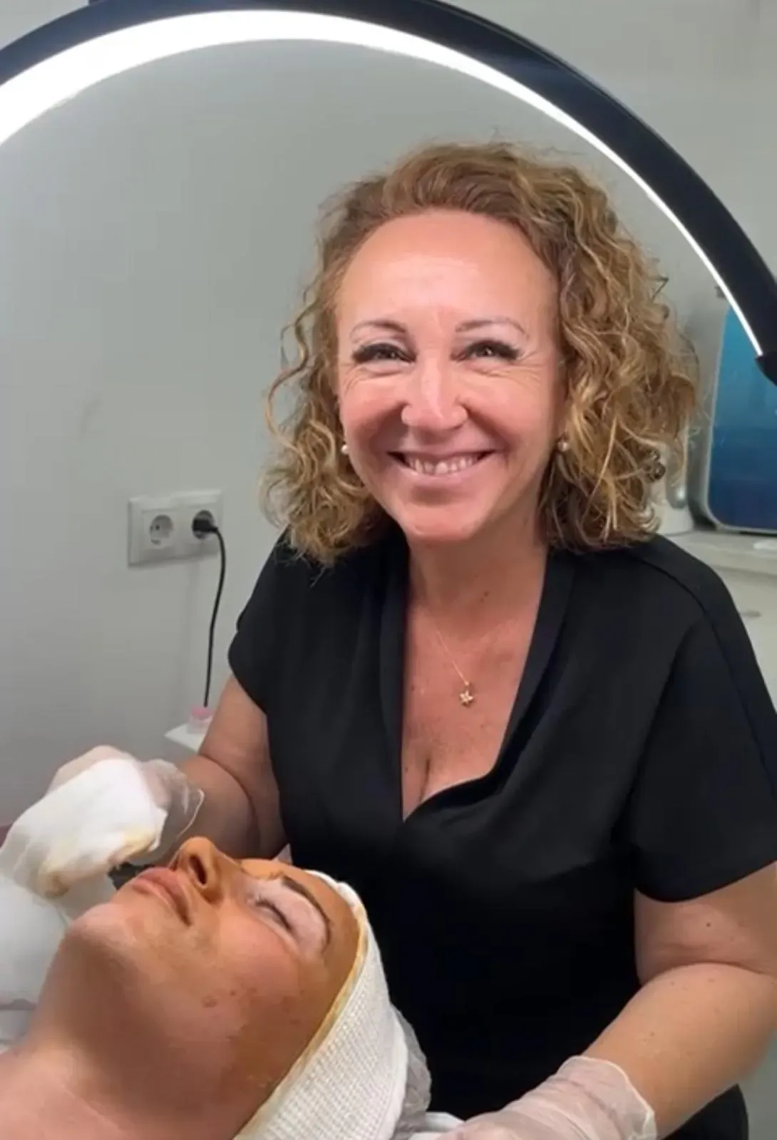 Una profesional con una blusa negra sonríe mientras realiza un tratamiento facial a una clienta en un entorno clínico.