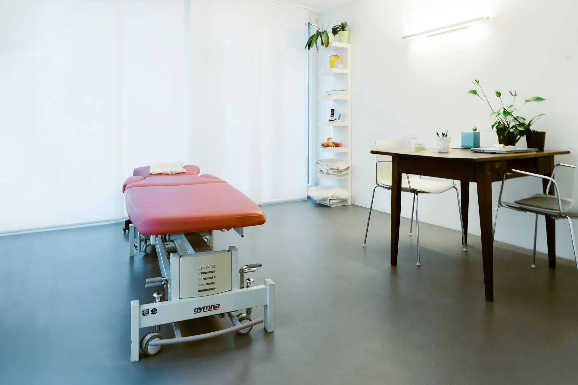 Leerer Behandlungsraum mit roter Massageliege, Holztisch, Stuhl und weissen Wänden