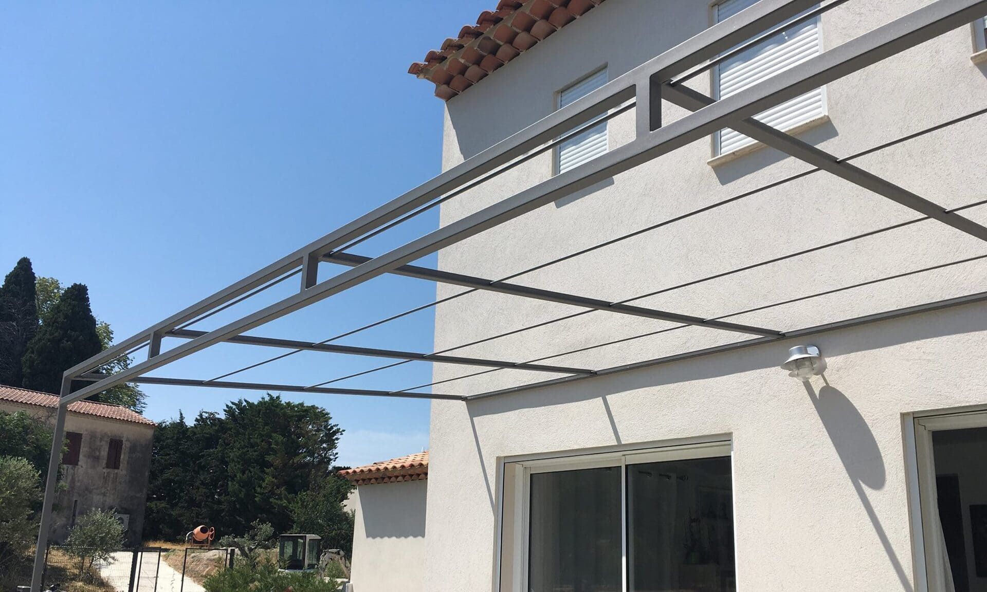 Pergola métallique adossée à une maison blanche, au-dessus d'une porte coulissante en verre, sur fond de ciel bleu.