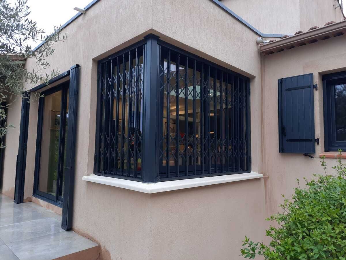 Extérieur d'une maison avec des barreaux de sécurité noirs sur une baie vitrée, volets noirs.