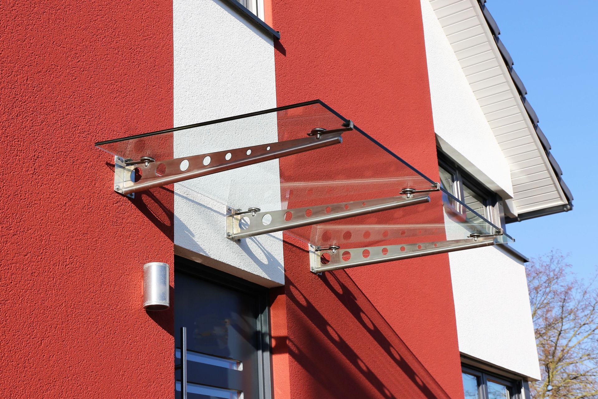 Auvent en verre au-dessus d'une porte, fixé à la façade rouge et blanche d'un bâtiment.