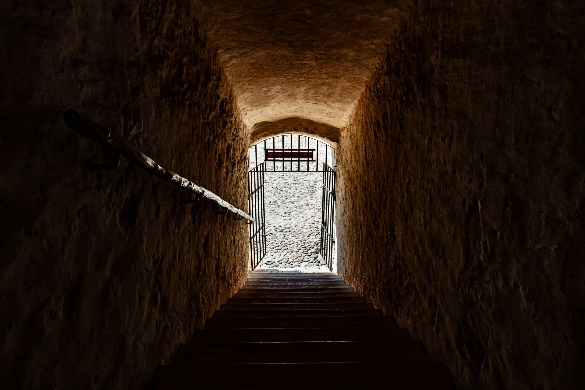 Tunnel en pierre avec escaliers descendants menant à une ouverture fermée par une grille, lumière vive au bout.