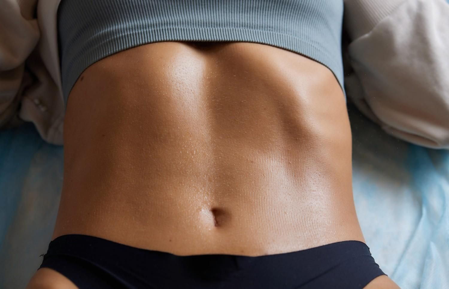 Primer plano de un torso desnudo con el ombligo visible, vistiendo pantalones cortos oscuros sobre una cama.