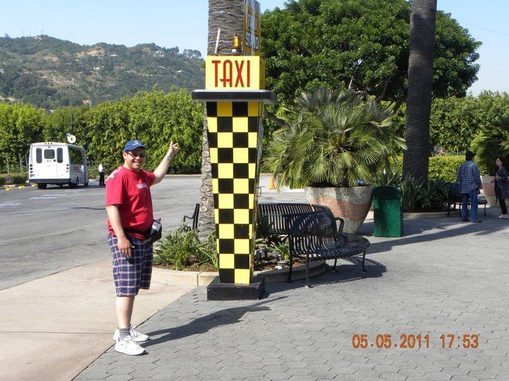 Una persona señala una señal de taxi. La señal es de cuadros negros y amarillos. Al fondo se ven una furgoneta, árboles y una montaña.