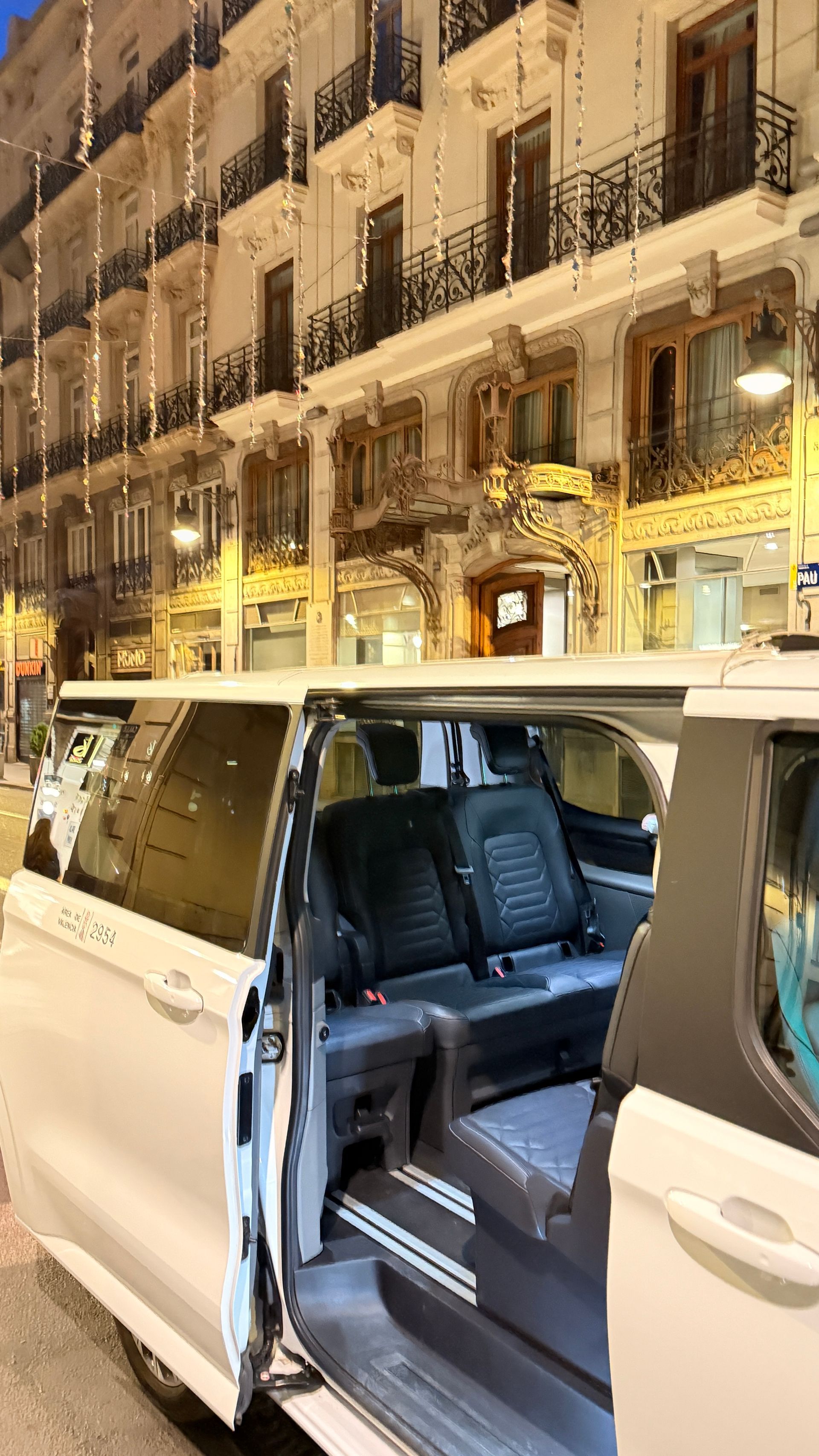 Una camioneta blanca estacionada en una calle de la ciudad, con la puerta abierta, revelando asientos negros. Edificios con balcones iluminados al fondo.