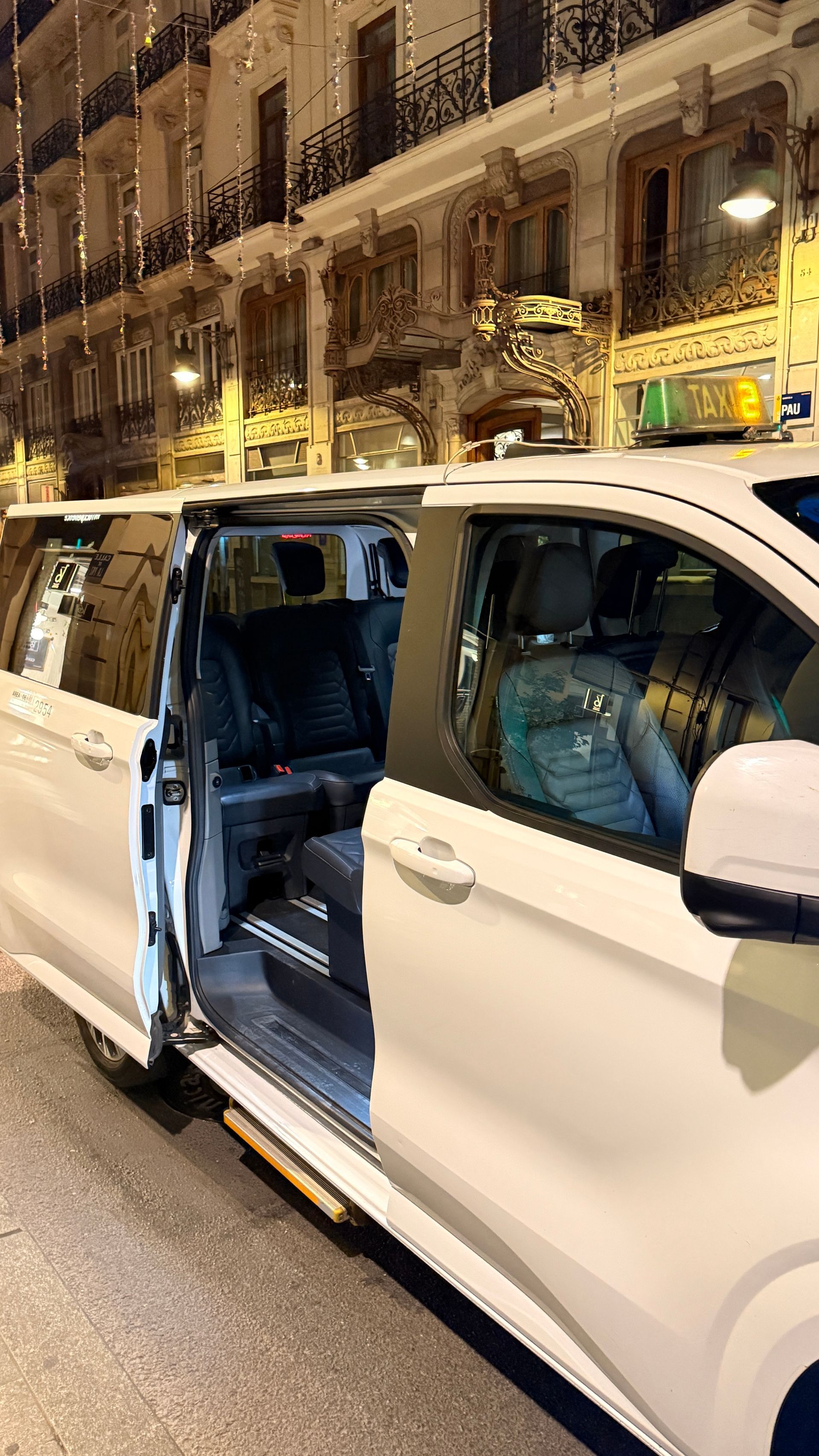 Taxi blanco estacionado en una calle de la ciudad por la noche, con la puerta abierta y el conductor visible.