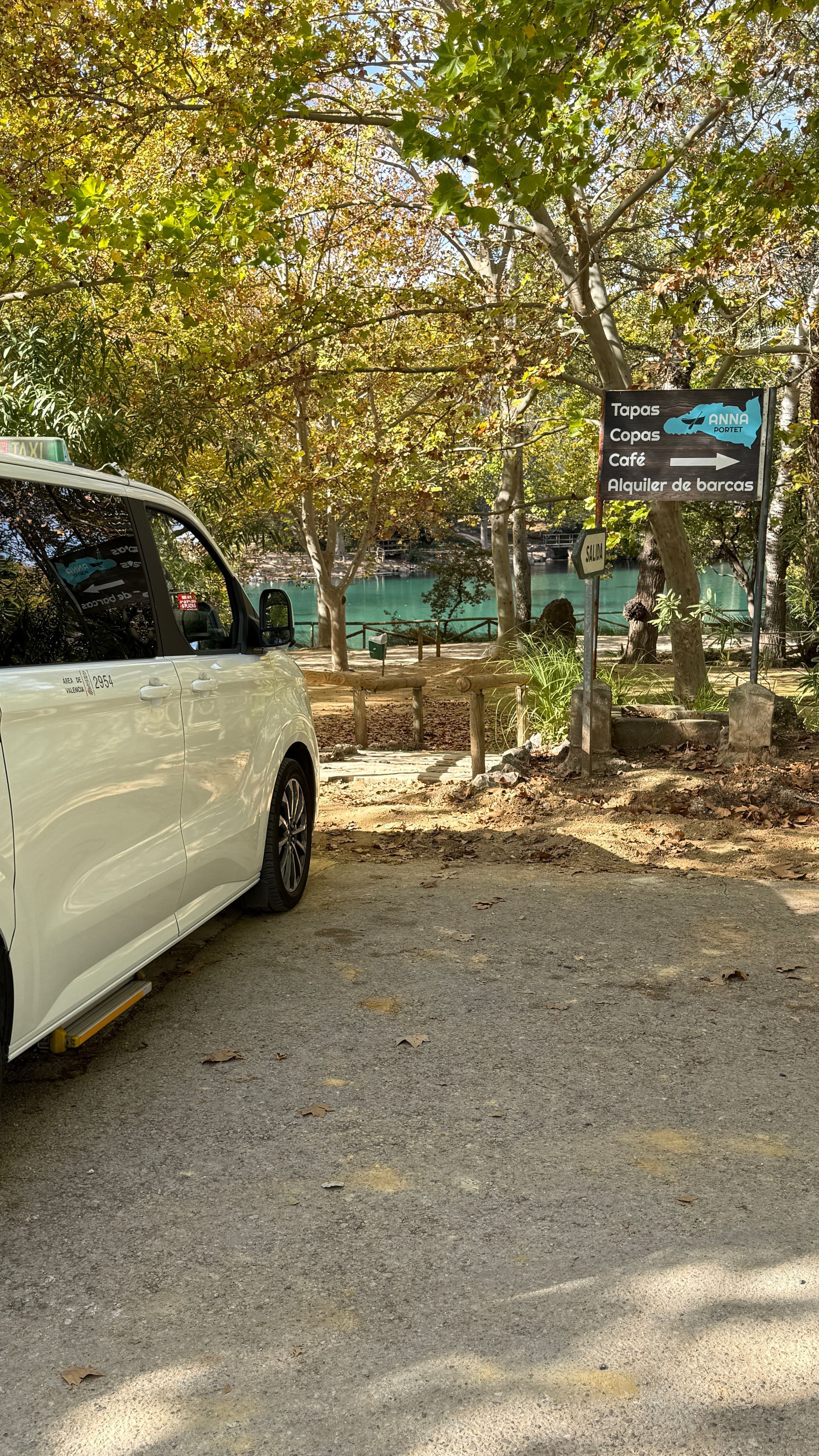Vehículo blanco estacionado sobre grava cerca de árboles, letrero y agua. Luz solar.