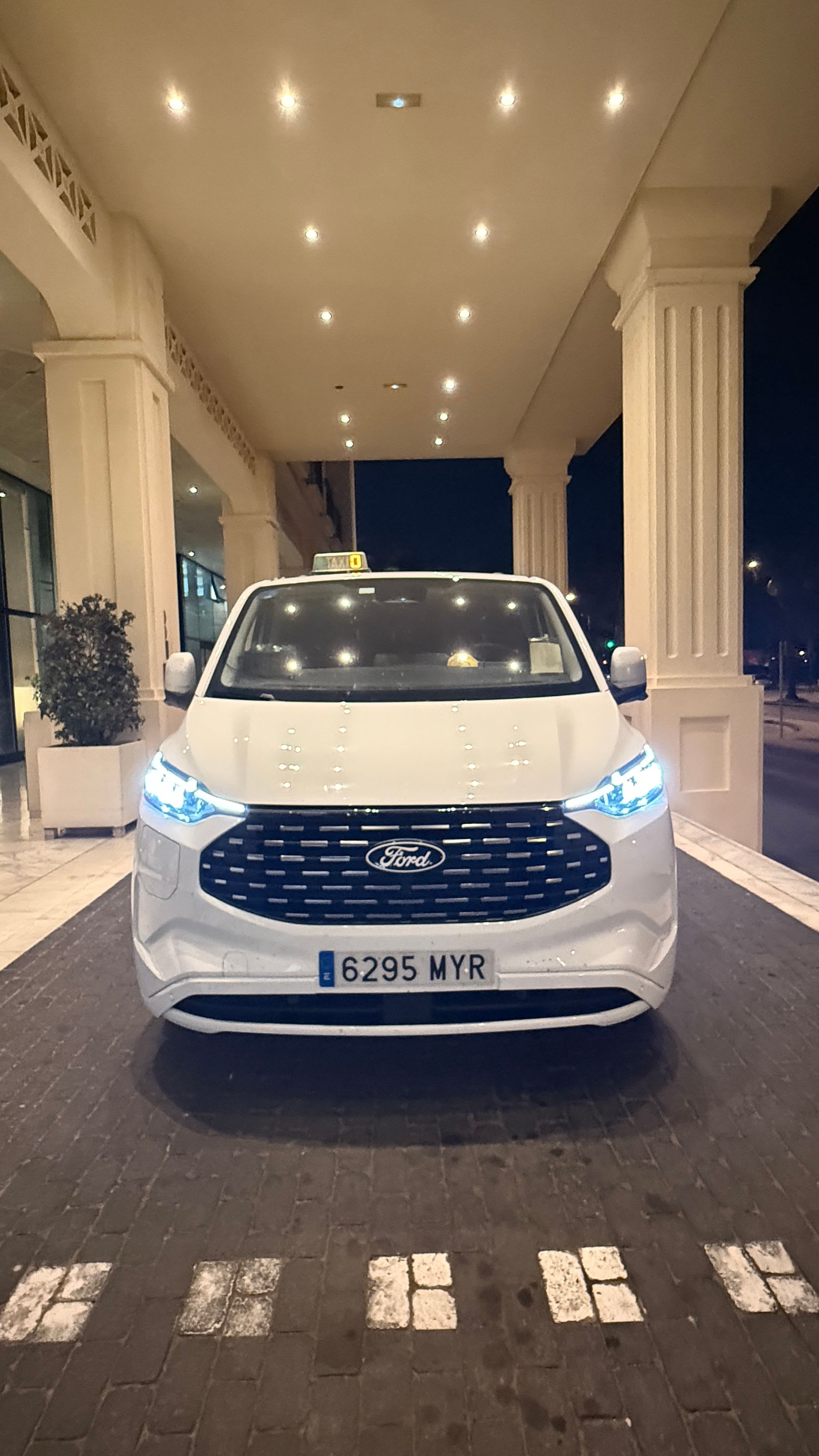 Taxi blanco aparcado bajo el toldo de un hotel de noche. Faros encendidos y matrícula visible.