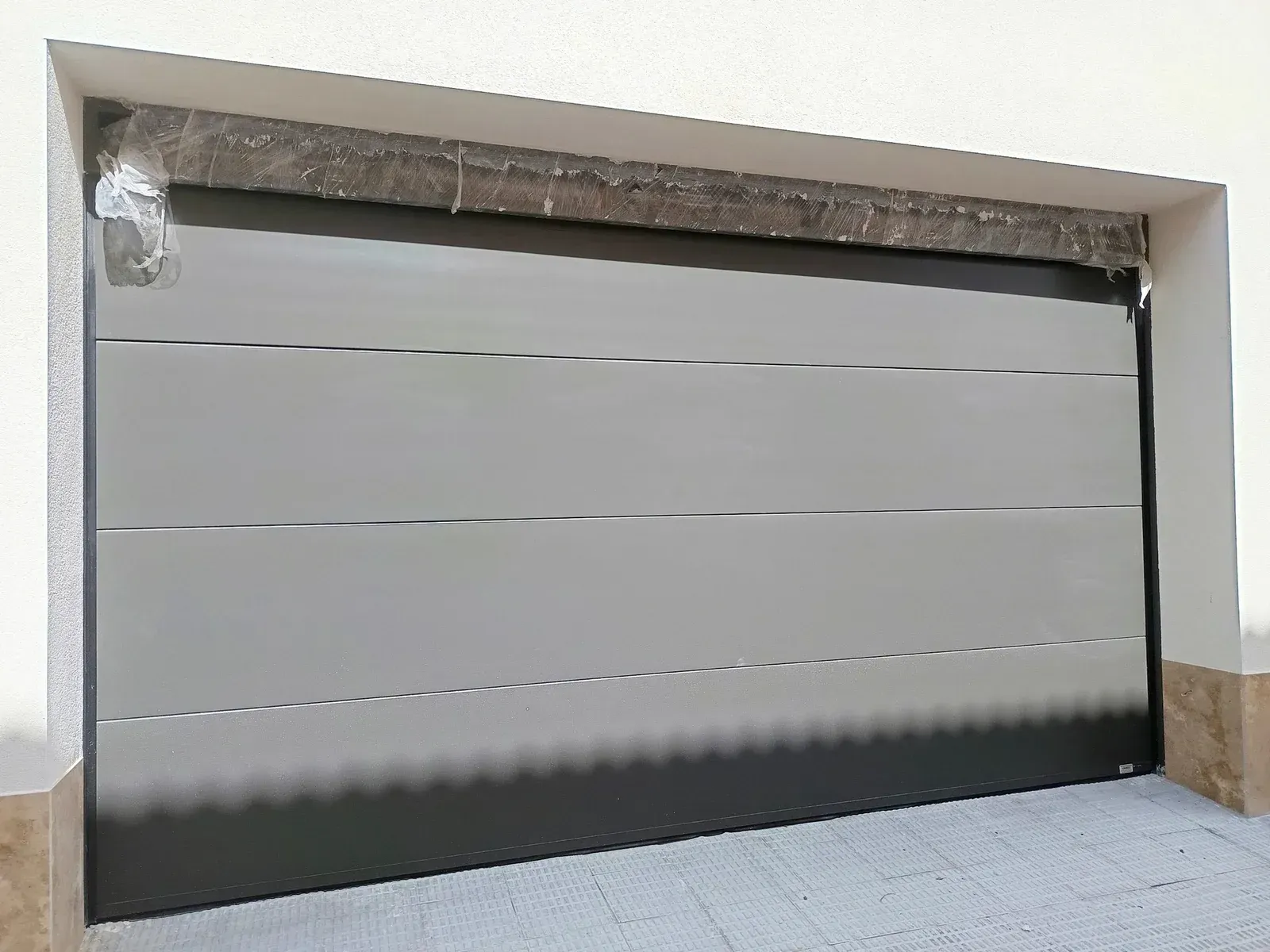 Puerta de garaje de color gris y negro con líneas horizontales, en exterior blanco y marrón.