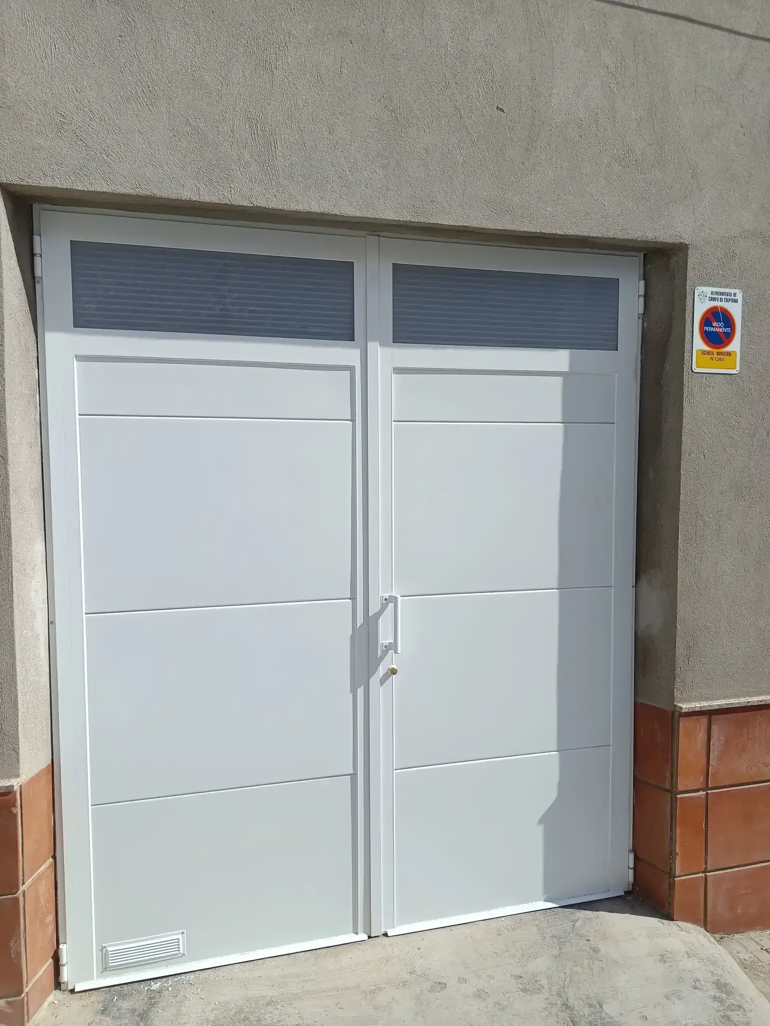 Puertas de garaje dobles blancas con paneles de vidrio esmerilado, colocadas en una pared de hormigón y un cartel de estacionamiento.