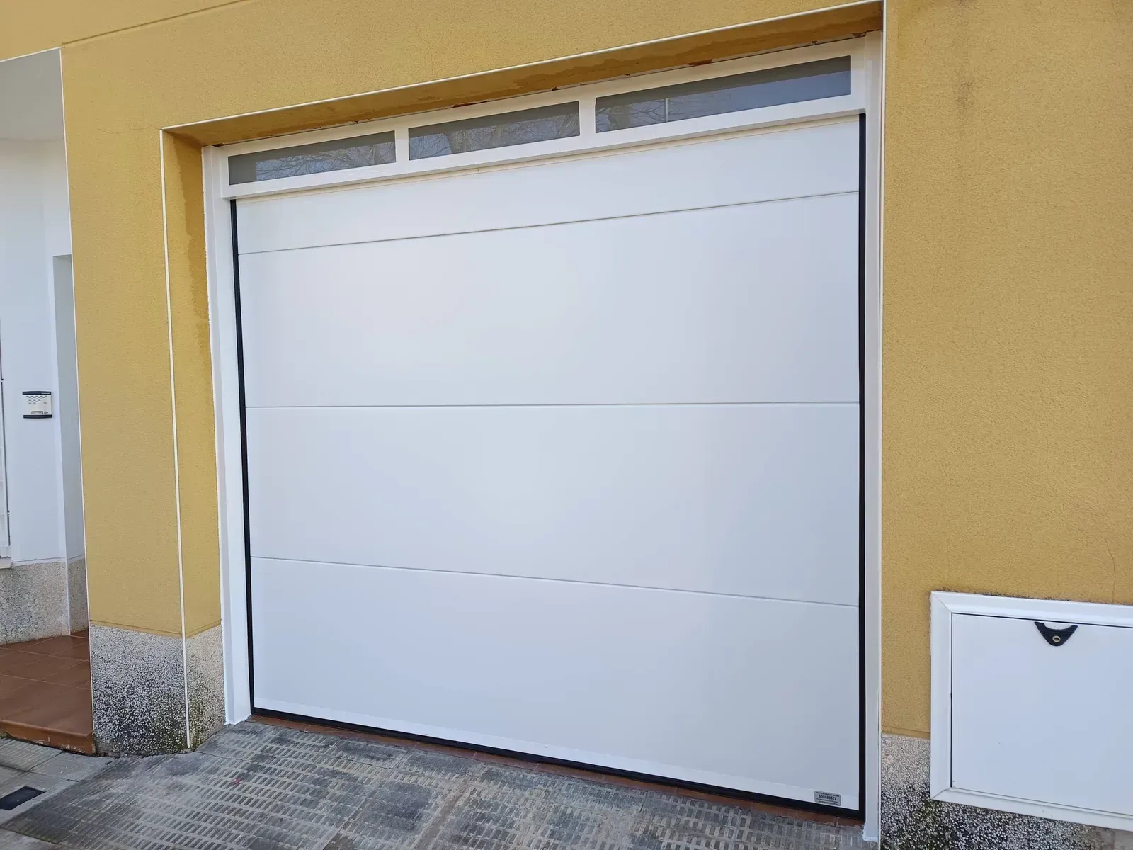Puerta de garaje blanca con tres paneles horizontales y una ventana rectangular en la parte superior, ubicada en un edificio amarillo.