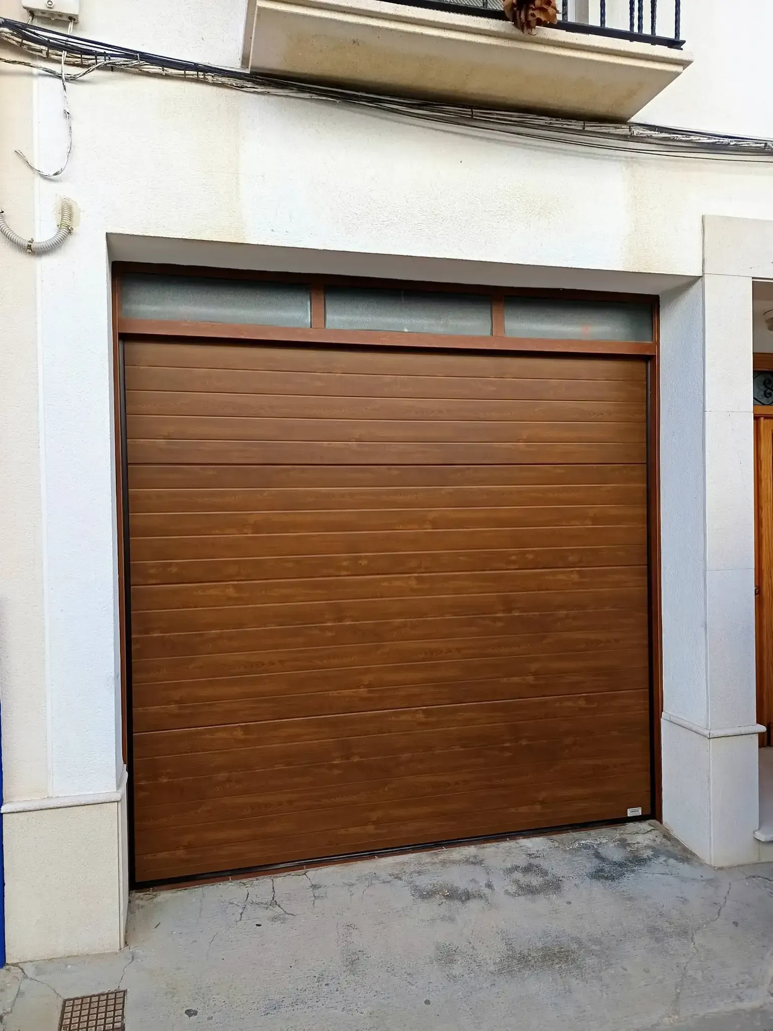 Puerta de garaje de color marrón con lineas horizontales, situada en una fachada de edificio de color blanco.