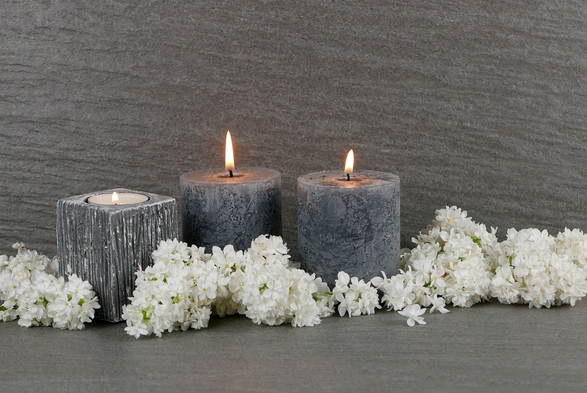 Drei brennende Kerzen und weiße Blumen auf einem Tisch.