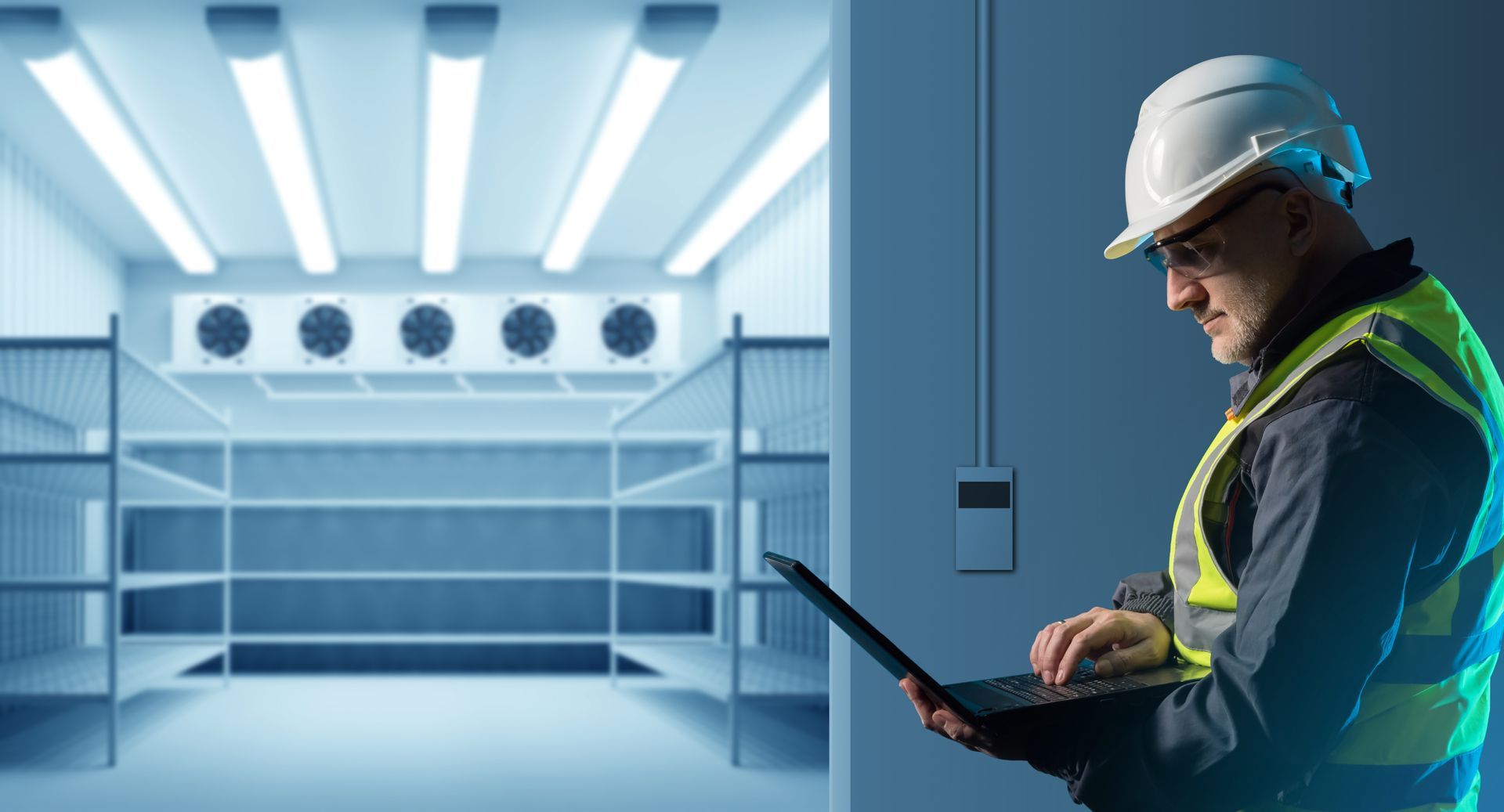 Hombre con casco y chaleco usando una computadora portátil frente a una cámara frigorífica.