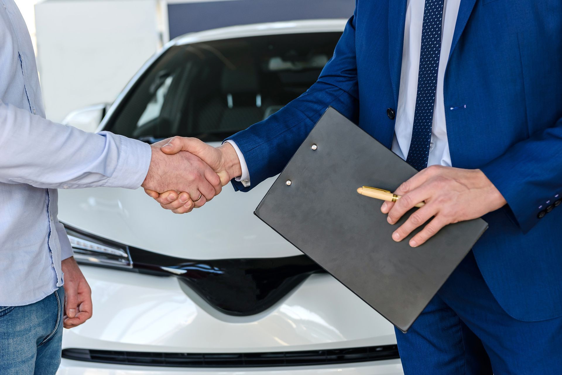 Un hombre con traje azul le da la mano a otro hombre delante de un coche blanco.