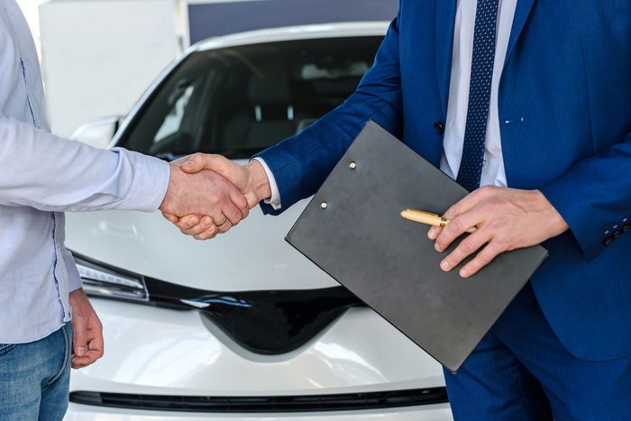Un hombre con traje azul le da la mano a otro hombre delante de un coche blanco.