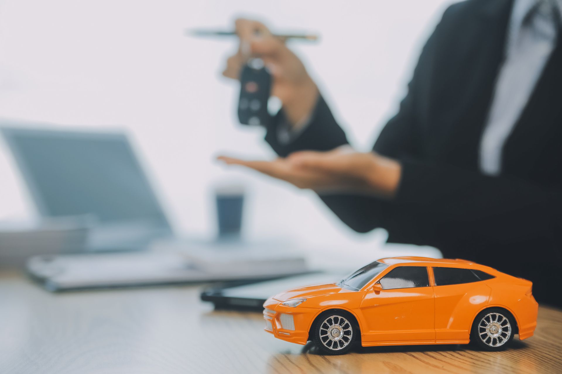 Persona con traje entregando las llaves del coche; coche de juguete naranja sobre un escritorio con una computadora portátil.