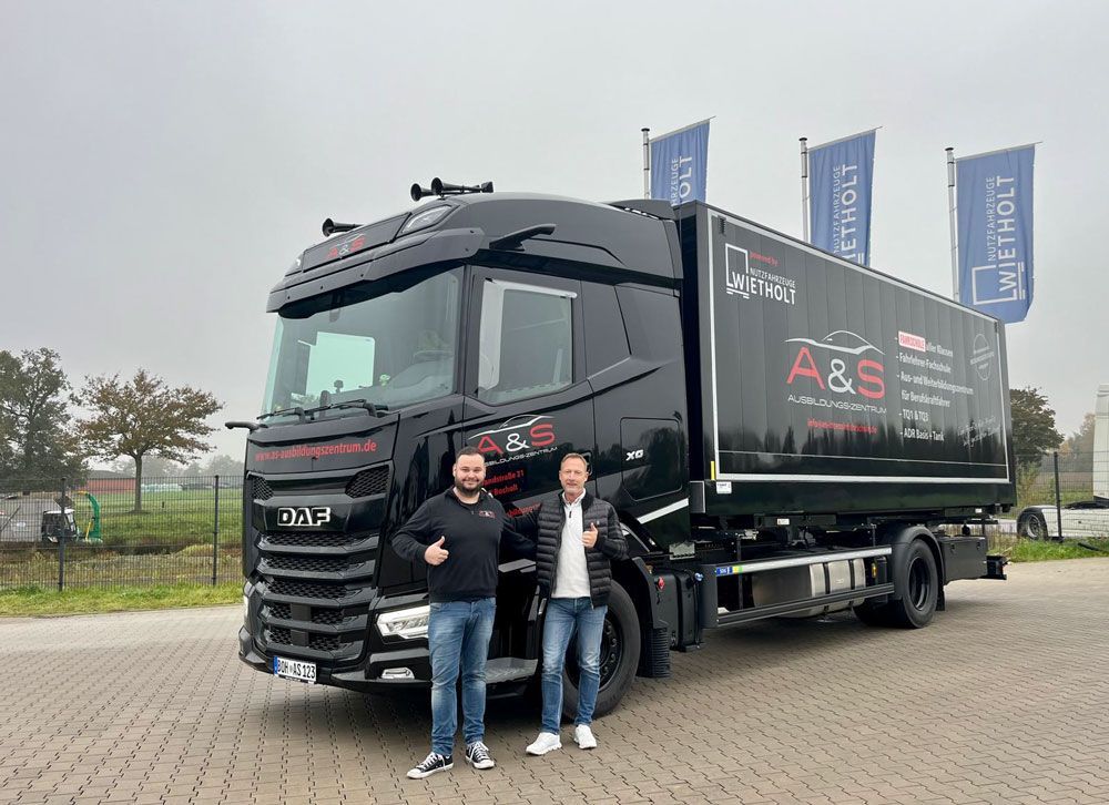 Zwei Männer stehen neben einem schwarzen Truck mit A&S-Logos. Die Männer zeigen mit dem Daumen nach oben nach draußen. Hinter dem Truck hängen Fahnen mit dem Logo.