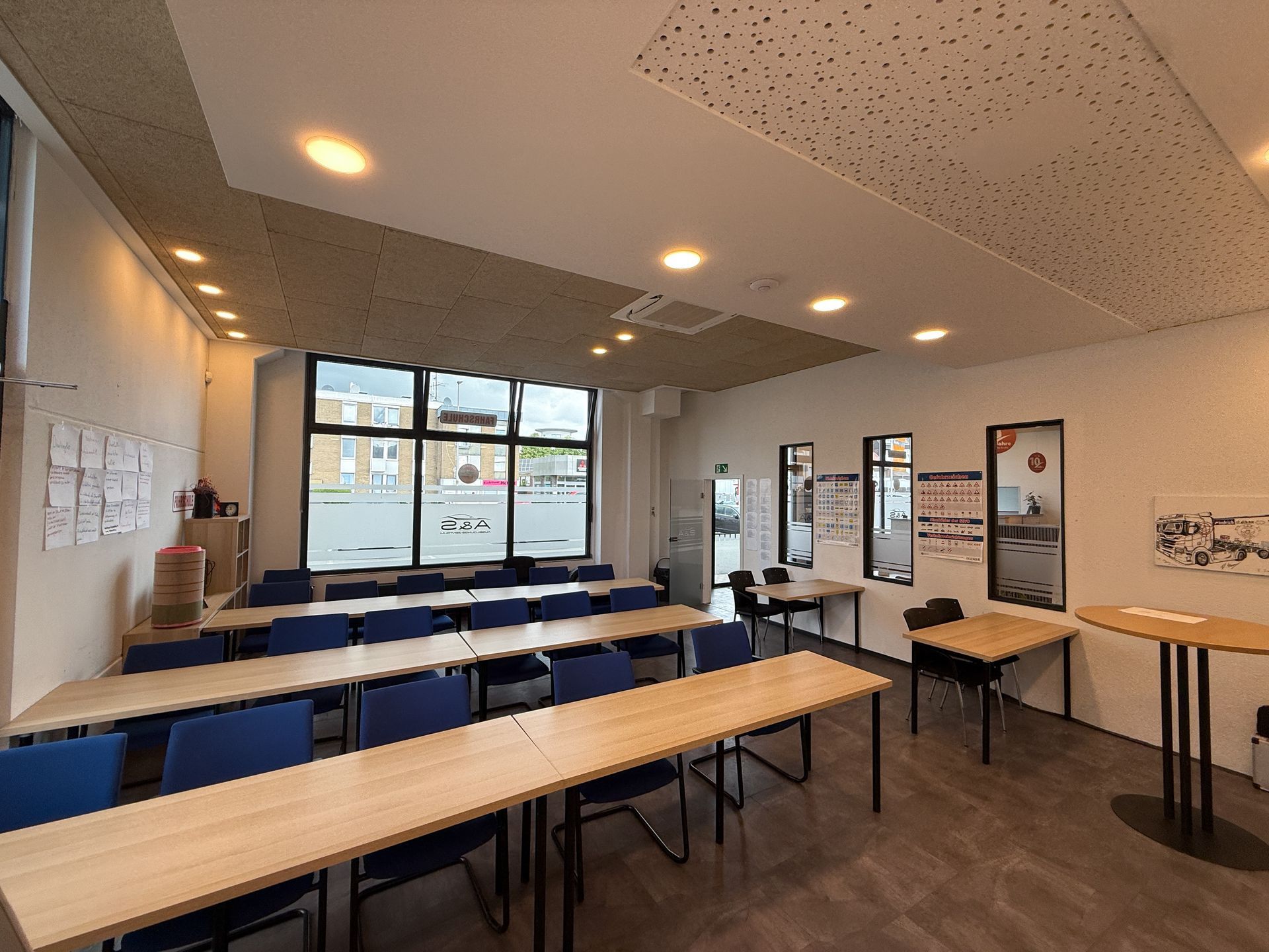 Klassenzimmer mit Reihen von Tischen, blauen Stühlen und einem großen Fenster mit Blick auf einen Parkplatz.
