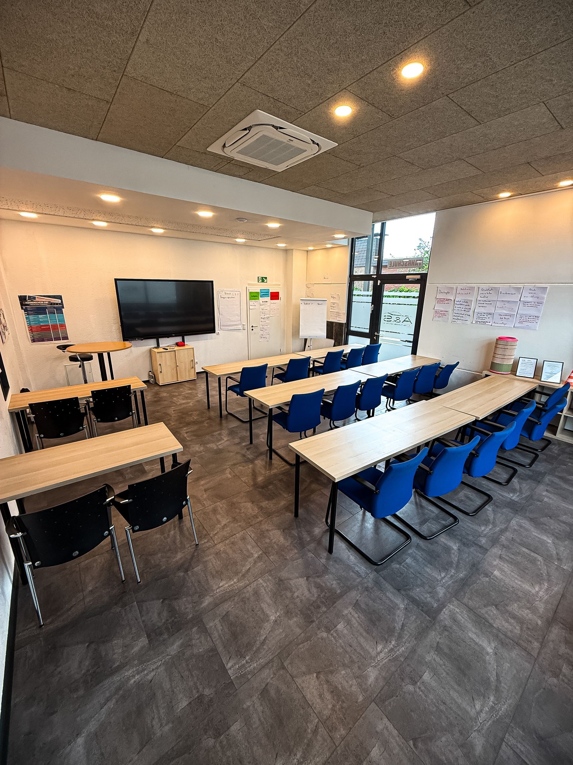 Klassenzimmer mit Reihen von Tischen und blauen Stühlen, weißen Wänden und einer großen schwarzen Leinwand.