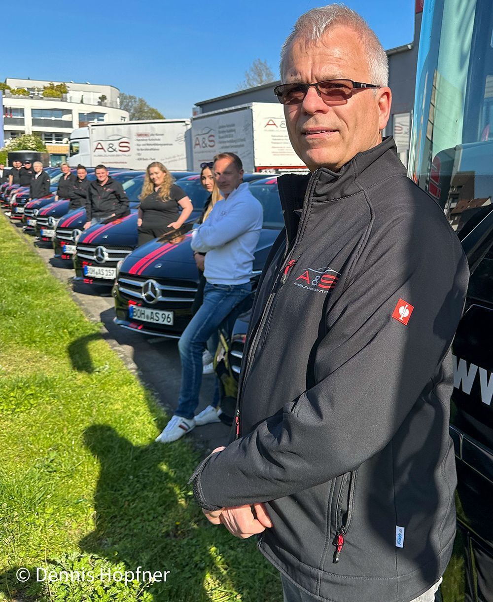 Ein Mann in einer schwarzen Jacke steht vor einer Reihe von Autos.