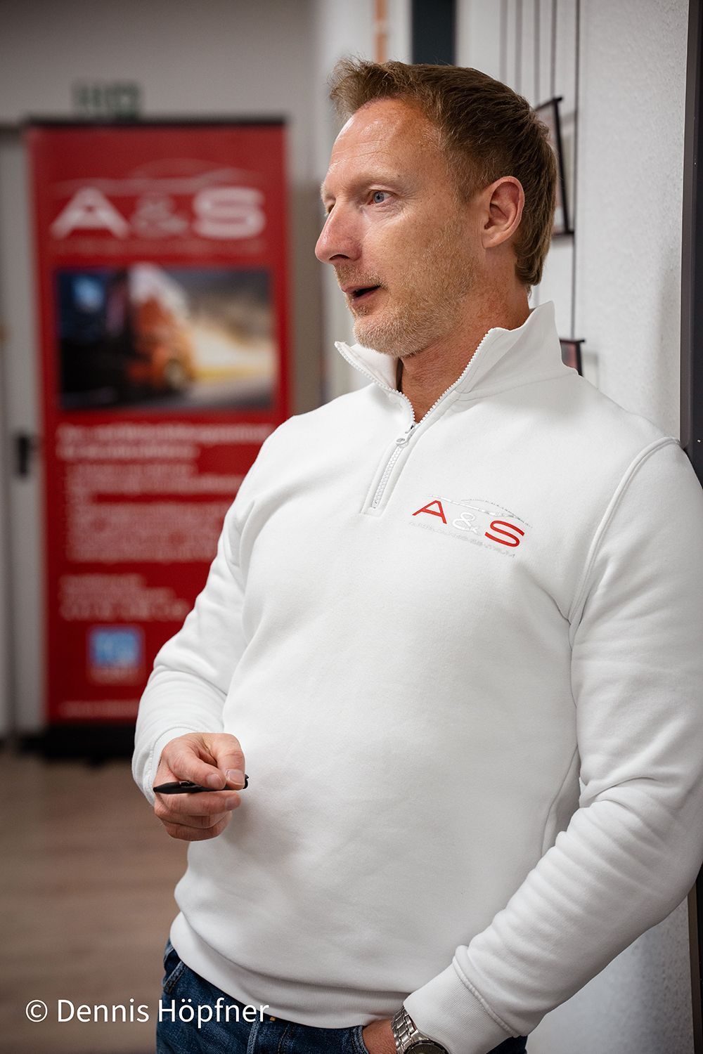 Ein Mann in einem weißen Pullover steht vor einem roten Schild.