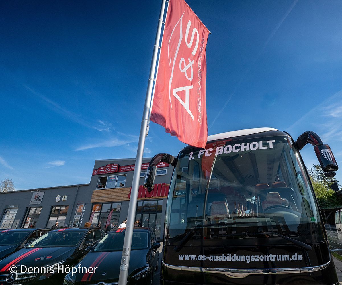 Ein schwarzer Mannschaftsbus mit einer roten Flagge mit der Aufschrift „A&S“ und einem Gebäude mit davor geparkten Autos.